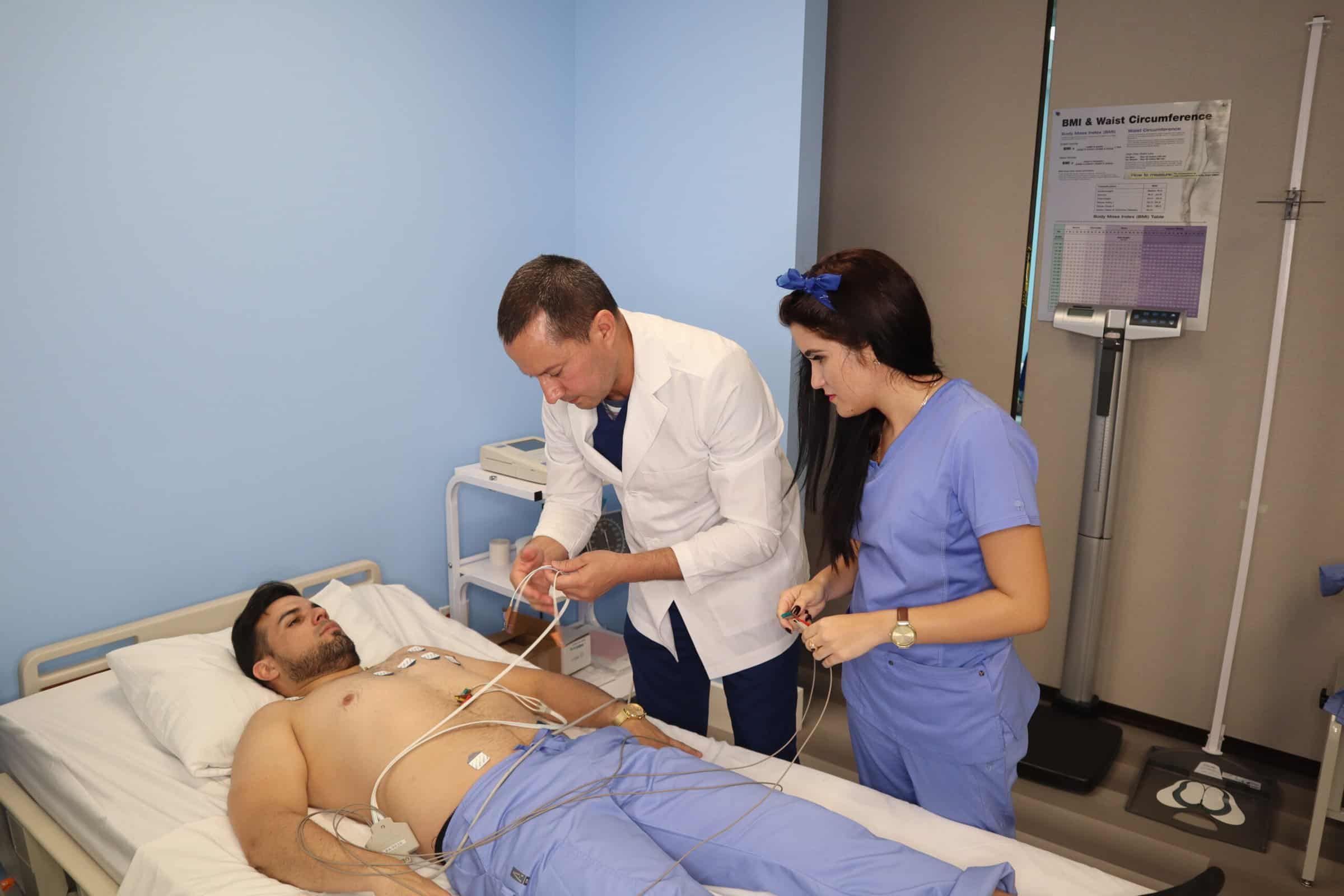 An instructor assists two Miami medical assistant training program students practicing in a skills lab, one is lying in a hospital bed while the other student stands next to the instructor and observes